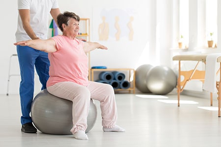 Elderly Senior Trying to Maintain Balance While Sitting on a Grey Fitball While Being Supported by Physical Therapist